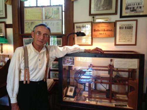 A man in a white shirt and suspenders stands beside a display case containing a historical model in a museum setting.