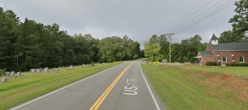 A quiet road lined with trees, leading to a church and a cemetery on the side. Overcast sky above.