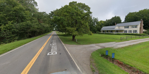 A quiet road named Broad Road lined with trees, leading to a large white house on a grassy lawn.