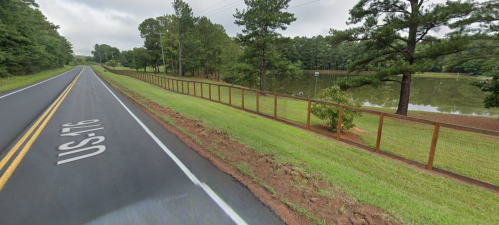 A quiet road lined with a fence, next to a calm pond surrounded by trees under a cloudy sky.