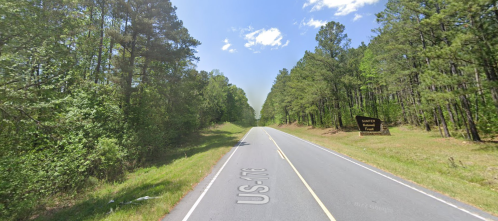 A two-lane road lined with trees under a clear blue sky, with a sign for "Tanner's Trail" visible on the right.