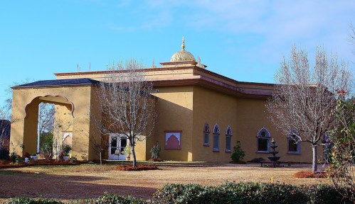 A yellow building with a dome and arched windows, surrounded by bare trees and a grassy area under a blue sky.