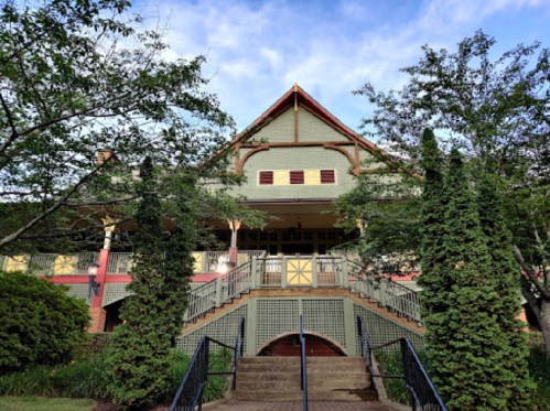 A large, two-story building with a peaked roof, surrounded by trees and greenery, featuring a staircase leading to the entrance.