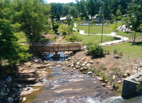 A serene park scene featuring a small stream, a wooden bridge, and green trees with a playground in the background.