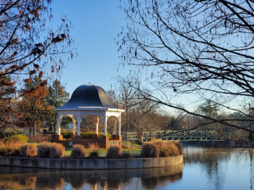 A gazebo by a calm pond, surrounded by trees and a bridge in a serene park setting.