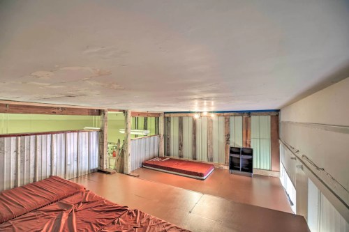 Loft space with a mattress on the floor, wooden walls, and a small window, illuminated by overhead lights.