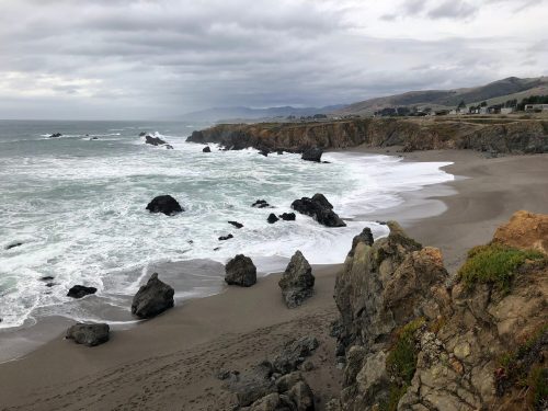 A rocky coastline with crashing waves under a cloudy sky, featuring sandy beaches and distant hills.