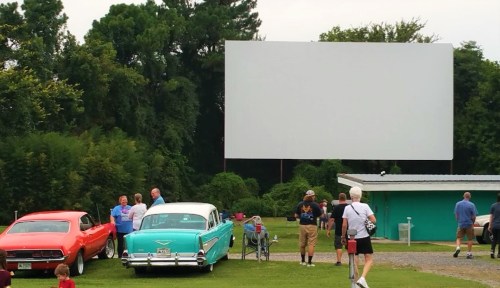 A drive-in movie theater scene with classic cars and people gathered in front of a large blank screen.