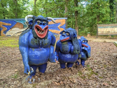 Three whimsical blue sculptures with exaggerated features stand in a wooded area, surrounded by fallen leaves and colorful murals.