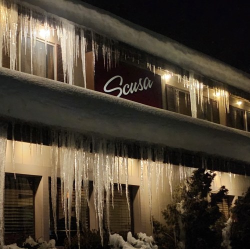 Icicles hang from a snow-covered roof at night, with a sign reading "Scusa" illuminated by warm lights.