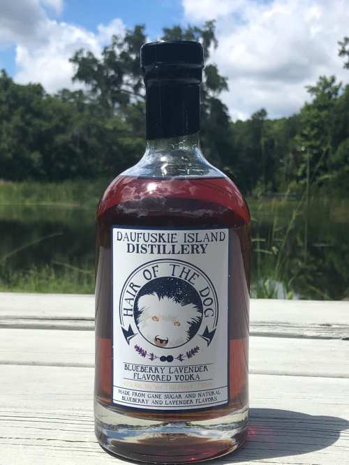 Bottle of Hair of the Dog blueberry lavender flavored vodka on a wooden surface, with a scenic background of trees and water.