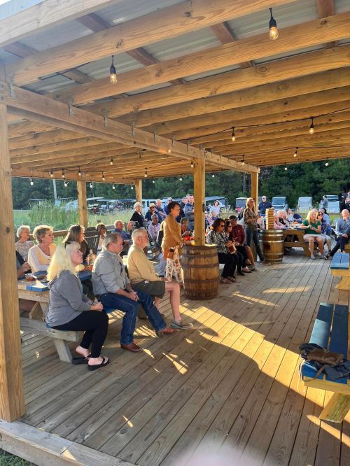 A crowd of people seated under a wooden pavilion, enjoying an outdoor gathering with string lights overhead.