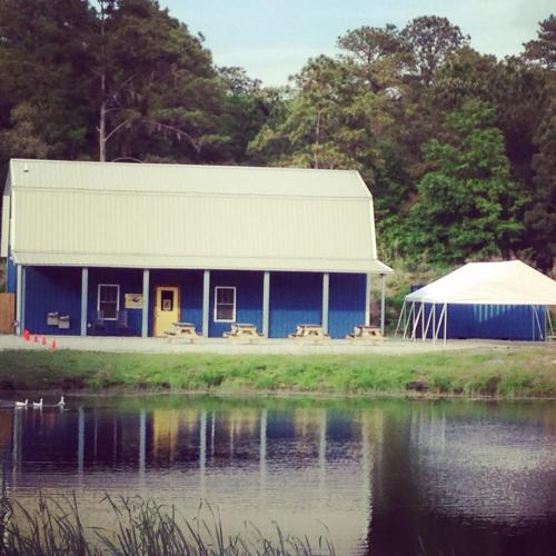 A blue building with a metal roof beside a pond, featuring picnic tables and a white tent nearby.