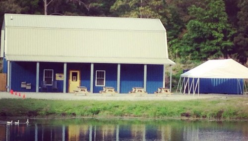 A blue building with a metal roof and picnic tables in front, near a pond and a white tent in the background.