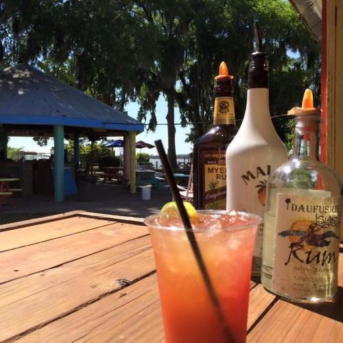 A tropical cocktail sits on a wooden table, with rum bottles and a scenic outdoor view in the background.