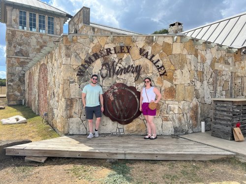 A couple stands in front of a stone winery sign, smiling on a sunny day. The building features rustic architecture.
