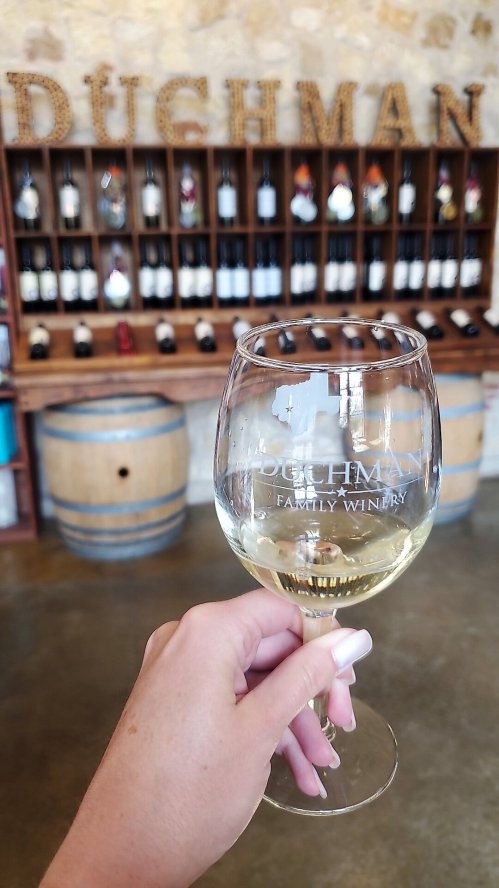 A hand holding a glass of white wine in front of a wine rack at Dughman Family Winery.