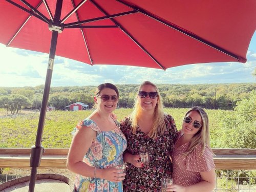 Three women stand under a red umbrella, smiling with drinks in hand, overlooking a vineyard and scenic landscape.