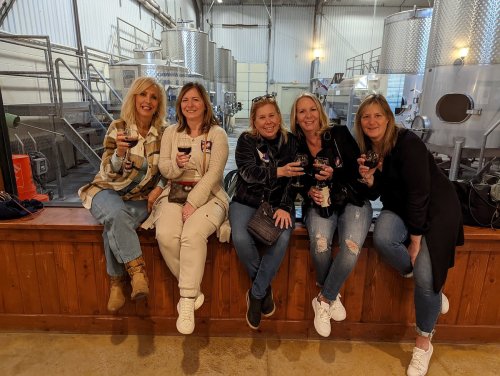 Five women holding drinks, smiling and sitting on a wooden ledge in a winery with stainless steel tanks in the background.