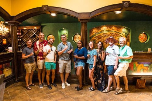 A group of nine people smiling and posing together in a colorful bar setting, holding drinks.
