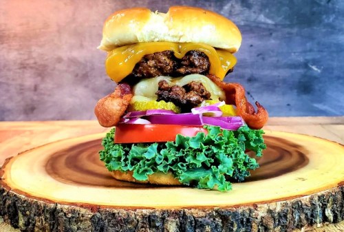 A towering burger with cheese, bacon, lettuce, tomato, pickles, and onions on a wooden platter.