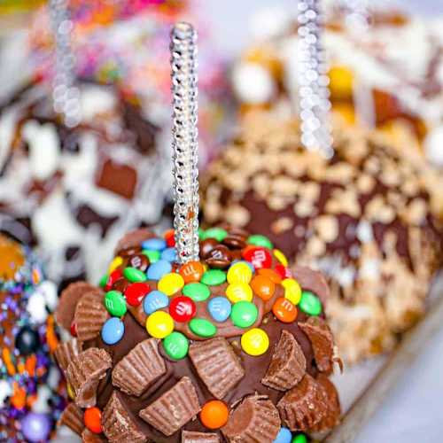 Close-up of chocolate-covered caramel apples topped with colorful candies and nuts, featuring shiny silver sticks.