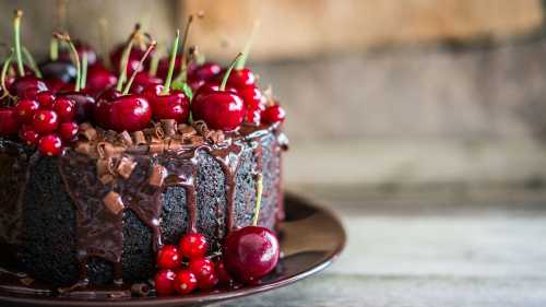 A rich chocolate cake topped with fresh cherries and red currants, drizzled with chocolate, on a wooden surface.