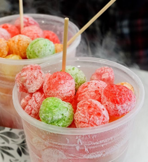 Two clear cups filled with colorful, frozen fruit balls in red, green, and orange, with wooden sticks for serving.