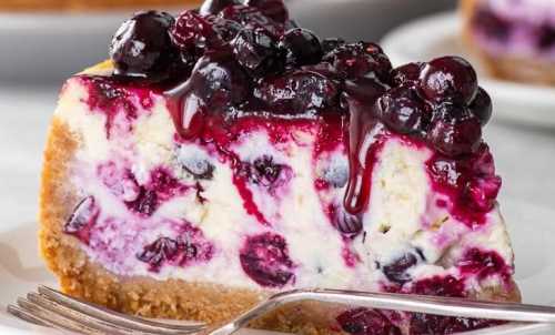 A slice of cheesecake topped with a glossy berry compote, served on a white plate with a fork beside it.