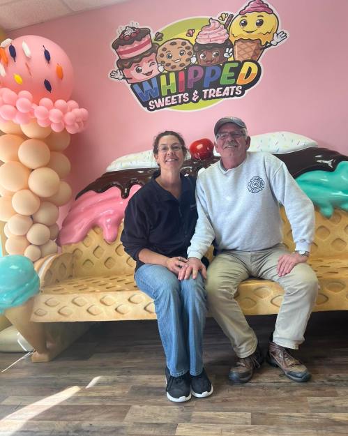 A couple sits on a colorful dessert-themed couch in a sweet shop, with a playful wall mural behind them.