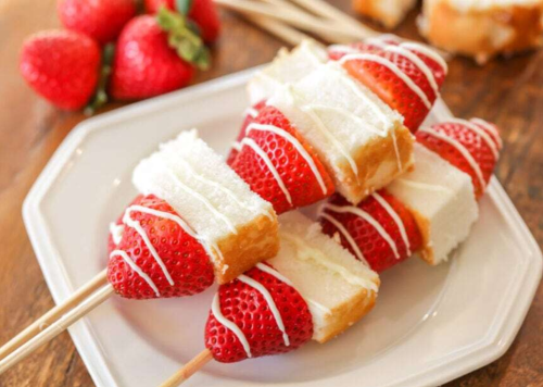 Skewers of strawberries and cake pieces drizzled with white chocolate, served on a white plate.