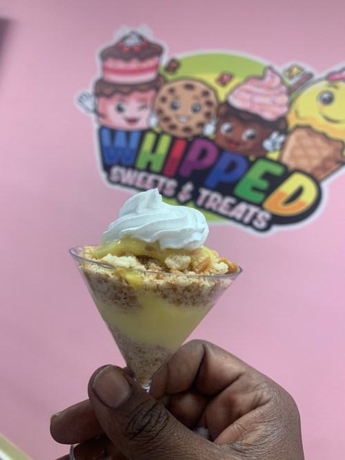 A hand holding a dessert cup with layers of cream and cake, in front of a colorful "Whipped Sweets & Treats" sign.