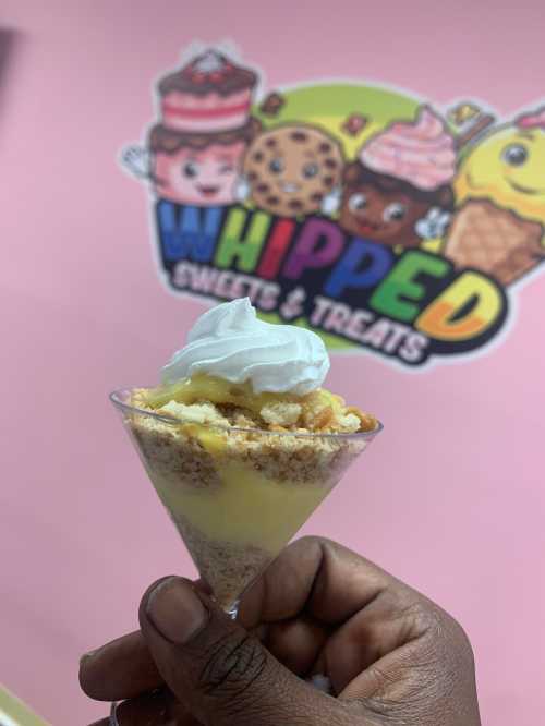 A hand holding a dessert cup with layers of cream and cake, in front of a colorful "Whipped Sweets & Treats" sign.