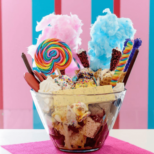 A colorful dessert bowl filled with ice cream, candy, cotton candy, and various sweet toppings against a striped background.