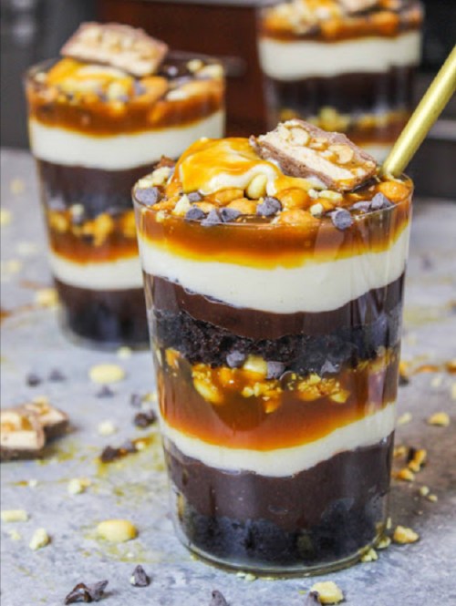 Layered dessert in a glass with chocolate, caramel, and crushed candy bars, topped with a golden spoon.