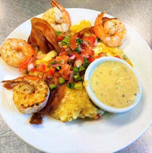 A plate of shrimp and grits topped with bacon, fresh pico de gallo, and a side of creamy sauce.