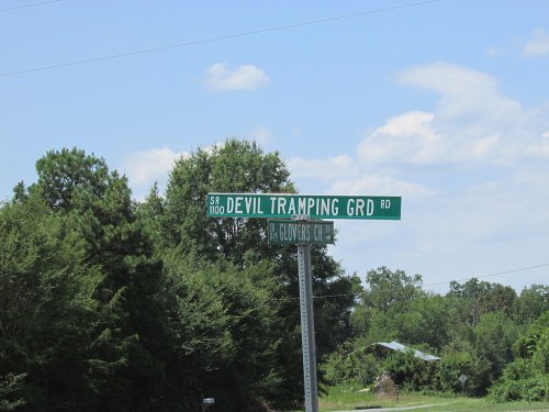 A green road sign reading "DEVIL TRAMPING GRD RD" against a backdrop of trees and a blue sky.