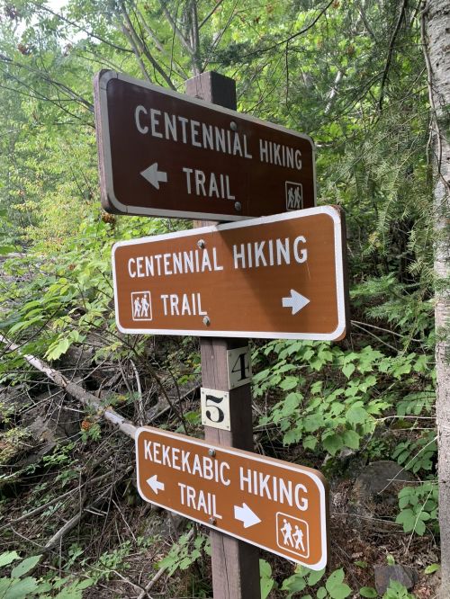 Three brown trail signs indicating directions for Centennial and Kekekabic Hiking Trails amidst greenery.
