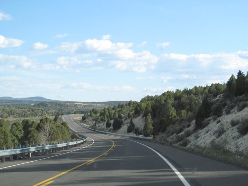 A winding road stretches through a landscape of trees and hills under a blue sky with scattered clouds.