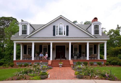 A charming gray house with a porch, surrounded by colorful flowers and greenery, featuring two people sitting outside.