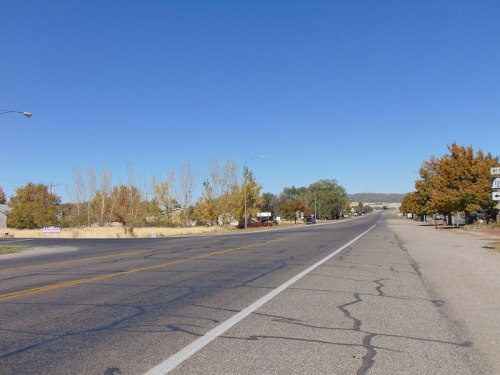 A wide, empty road lined with trees and buildings under a clear blue sky, showcasing autumn colors.