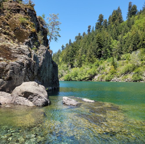 A serene river scene with clear water, rocky banks, and lush green trees under a bright blue sky.