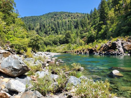 A serene river flows through a lush green landscape, surrounded by trees and rocky banks under a clear blue sky.
