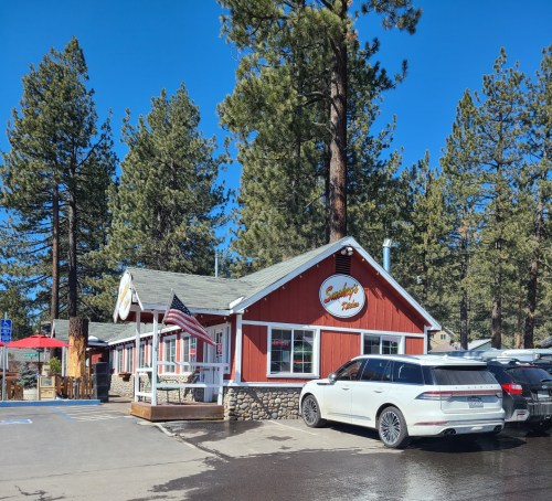 A cozy red and white restaurant surrounded by tall pine trees, with an American flag and parked cars in the foreground.