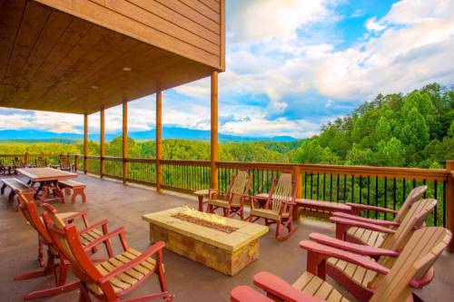 A spacious deck with wooden chairs and a fire pit, overlooking lush green mountains and a blue sky with clouds.