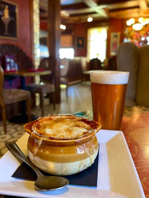 A bowl of French onion soup with melted cheese, served on a plate beside a pint of beer in a cozy restaurant setting.