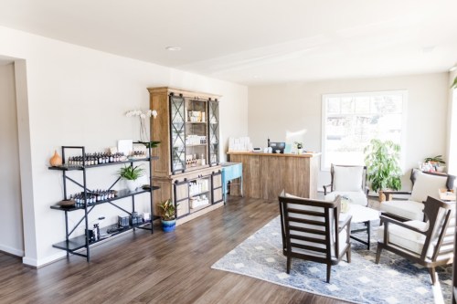 A bright, modern reception area with wooden furniture, plants, and shelves displaying bottles. Cozy seating is arranged.