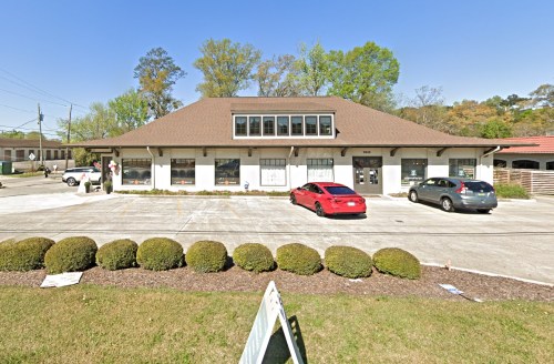 A single-story commercial building with a brown roof, surrounded by greenery and parked cars in a sunny setting.