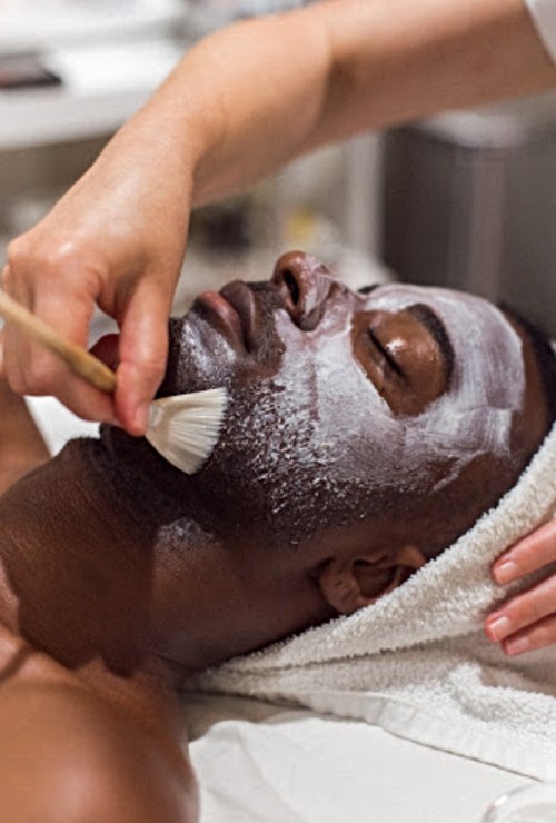 A person receiving a facial treatment, with a mask applied and a hand brushing on more product.