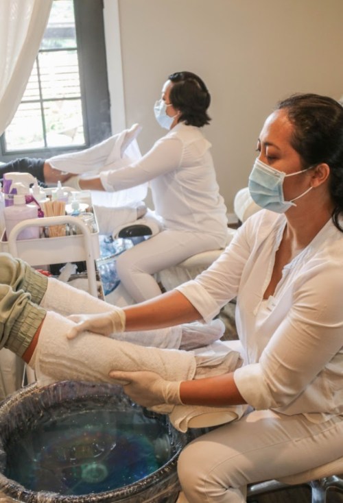 Two professionals in white attire provide spa treatments, wearing masks and gloves, in a serene indoor setting.